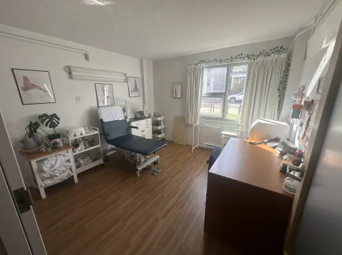 A bright and welcoming foot care clinic room featuring a blue adjustable treatment chair, anatomical foot posters on the wall, and organized medical supplies on nearby carts and shelves. A large window with white curtains lets in natural light, and a wooden desk with office essentials sits near the entrance, creating a clean and professional atmosphere.