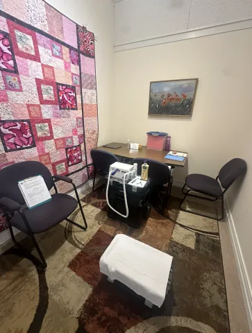 A cozy foot care clinic room with a patterned carpet, a white towel-covered footrest, and several chairs surrounding a wooden desk. The desk holds medical supplies, including a foot care device, lotion, and a sharps container. A colorful pink patchwork quilt hangs on one wall, and a framed painting of orange flowers decorates another, giving the space a warm and welcoming atmosphere.