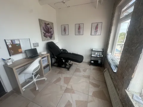 A clean and modern treatment room with a black reclining chair, a small wooden desk with a laptop and office supplies, anatomical foot posters on the wall, and a rolling cart with medical or skincare products. A large window lets in natural light, revealing a partial view of an outdoor area.