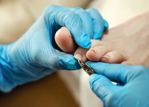 A close-up of a person wearing blue medical gloves carefully trimming another person's toenails with a metal nail clipper, focusing on foot care or a pedicure procedure.
