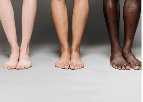 A neutral-gray background featuring three pairs of bare lower legs and feet—one pale, one tan, and one dark-skinned—standing side by side, toes forward.