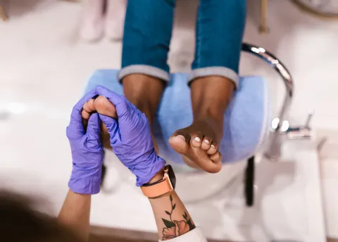 A person wearing purple gloves gently holds and massages the foot of another individual seated with legs resting on a padded footrest, likely during a pedicure or reflexology session.